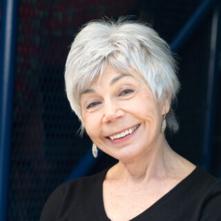 Portrait of poet Jane Munro, a white woman with short silver hair. She is wearing a black top and dangly earrings. She is leaning against a wall with her arms crossed, smiling at the camera.
