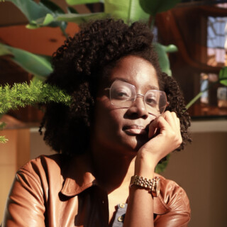 Portrait of poet Medgine Mathurin, a Black woman with medium-length dark hair and wire rim glasses. She is wearing a reddish brown leather jacket and is leaning on a glass countertop.