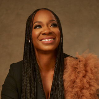 Portrait of poet Titilope Sonuga, a Black woman with long dark braided hair. She is wearng a black blouse with a brown ruffle. She is seated and smiling widely.