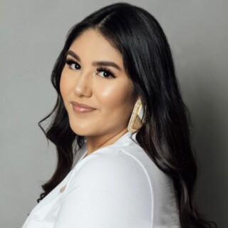 Headshot of poet Paige Cardinal, an Indigenous woman with long dark hair and dark eyes. She is wearing a white t-shirt and traditional earrings.