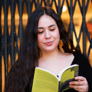 Headshot of poet Emily Riddle, an Indigenous woman with long dark wavy hair. She is reading from a book with a lime green and grey cover.