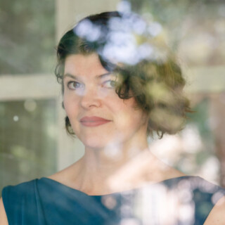 Portrait of poet Jennifer Bowering Delisle, a white woman with short dark wavy hair. She is wearing a teal sleeveless top.