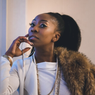 Portrait of poet Charlie Queen XO aka Charlene Smith, a Black woman with long dark hair in a high ponytail. She is wearing a white jacket, white crop top, and white pants with a faux fur wrap. She is leaning against a wall with her hand near her chin.