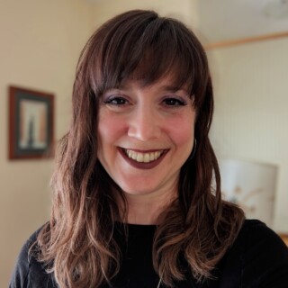 Headshot of poet Jessica Coles, a white woman with shoulder-length brown hair and brown eyes. She is smiling widely and wearing dark lipstick and a black top.