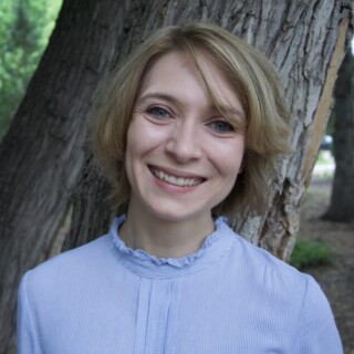 Headshot of poet Lauren Seal, a white woman with chin-length dark blonde hair. She is wearing a lilac blouse and is smiling. She is standing outside in front of a tree.
