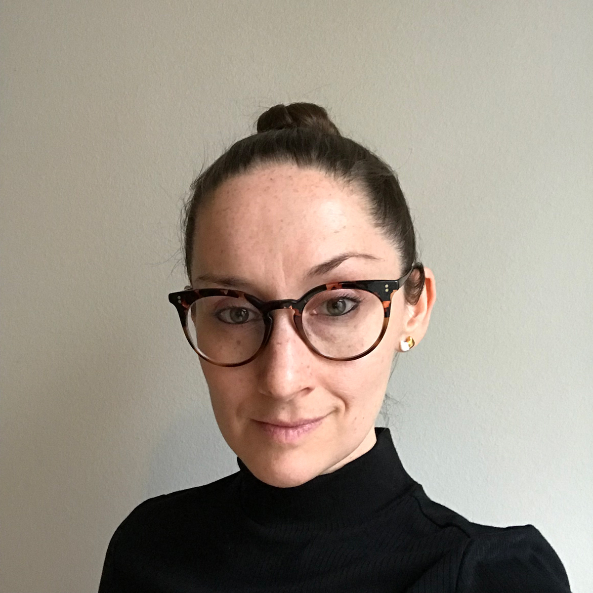 Headshot of M.L. Martin, a white non-binary poet with dark hair pulled back into a high bun wearing a black turtleneck and oversized glasses with black frames