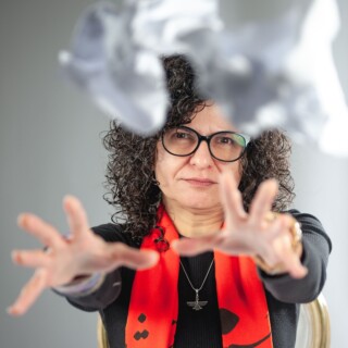 Portrait of poet Bänoo Zan, a white woman with curly shoulder-length dark hair and brown eyes. She is wearing a black long-sleeved shirt and a red scarf with a black printed design and glasses with black frames. She is throwing a crumpled up piece of paper at the camera.