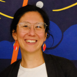 Portrait of poet Kathryn Gwun-Yeen Lennon, a woman of mixed white and east Asian heritage with dark hair pulled back into a ponytail and dark eyes. She is wearing wire-rimmed glasses, red earrings, a black blazer over a white t-shirt with a colourful print on it and jeans. She is in front of a wall mural and smiling at the camera.